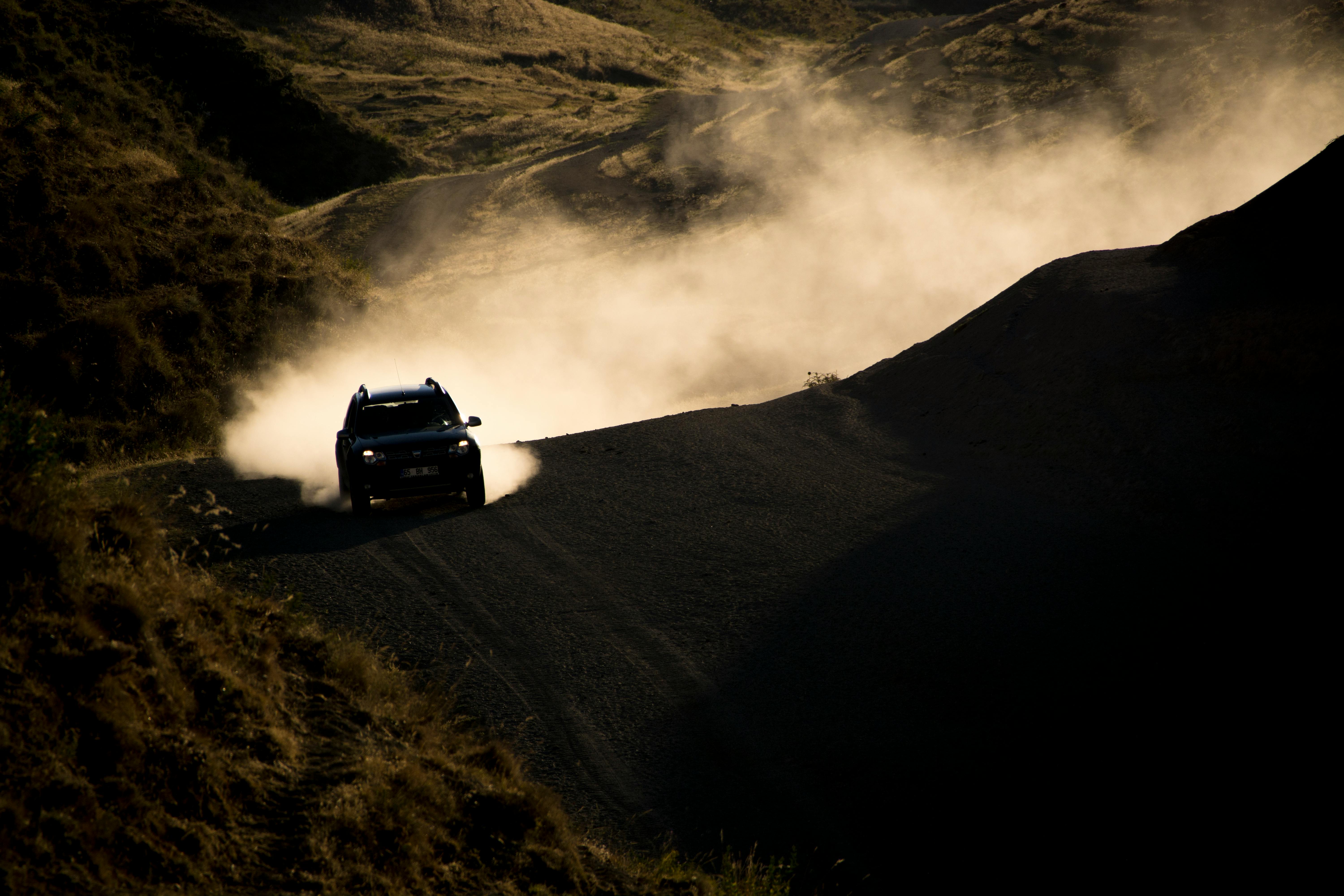 SUV driving fast on a dusty rural road, creating clouds of dust in the mountainous landscape.